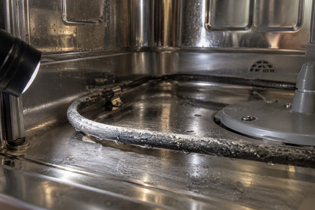 The dark curved heating element at the bottom of a dishwasher tub with a pale chalky crust along its surface.