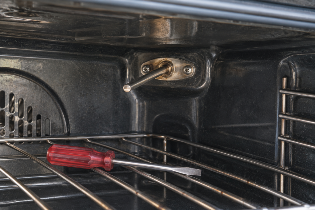 The straight metal temperature probe on the back wall of an empty oven cavity, held by two screws, with a screwdriver on the rack below.