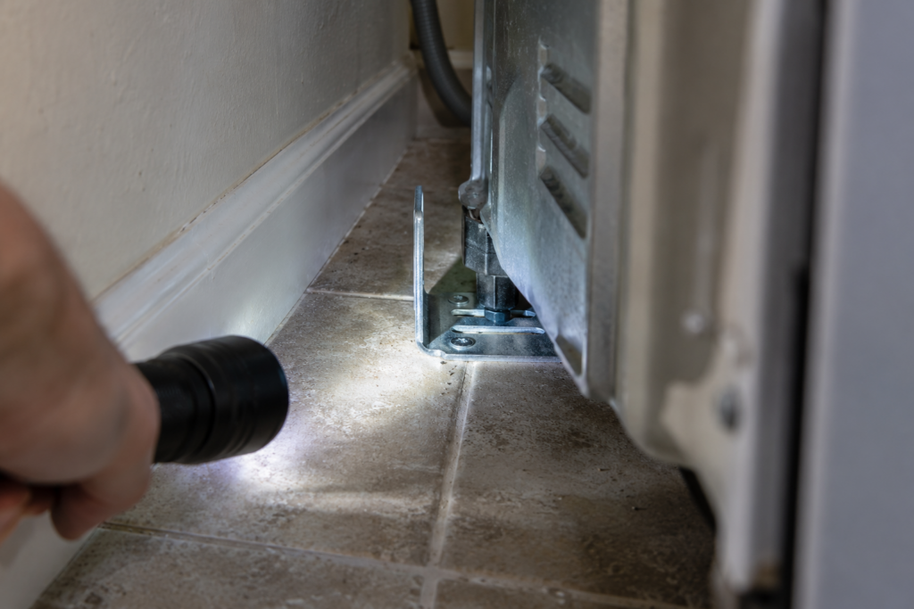 A flashlight beam on a steel anti-tip bracket screwed to the floor behind a range, with the rear leg seated in it.